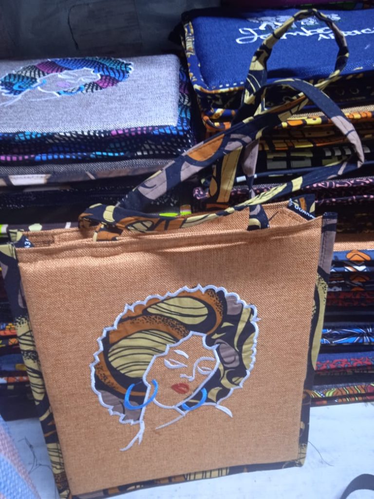 A beautiful celebration of heritage and style. This tan-colored bag features a stunning embroidered portrait of an African woman adorned in a vibrant headwrap and bold blue hoop earrings. Striking Artistry: A bold, artistic embroidered portrait that serves as a conversation starter. Natural Texture: Made with a burlap-textured fabric that offers a rustic yet sophisticated look. Coordinated Design: The patterned straps and side trim perfectly complement the colors in the central portrait. Unique Detail: Every bag features unique pattern variations on the handles, making each piece one-of-a-kind.